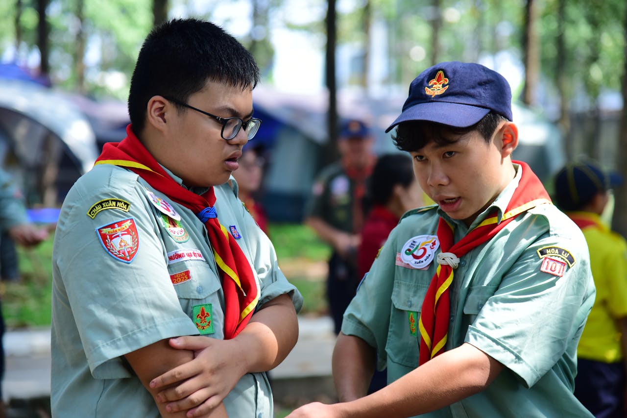 Two boy scouts in uniform interact during an outdoor camping session in a forested area.